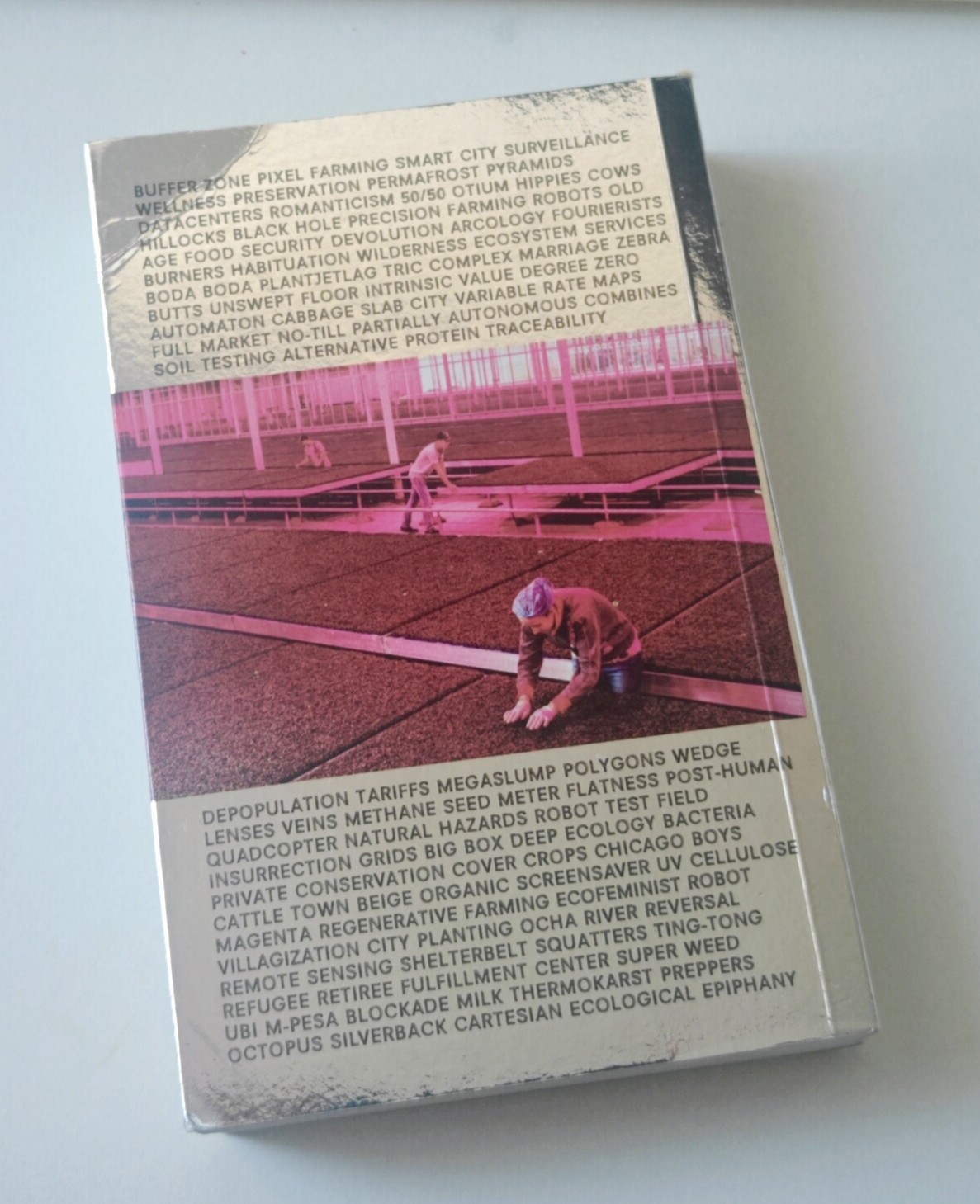 The back cover of Countryside: A Report — a pink-tinted photograph of workers tending rows of crops, surrounded by lists of the book's themes, from BUFFER ZONE and PIXEL FARMING to ECOLOGICAL EPIPHANY.
