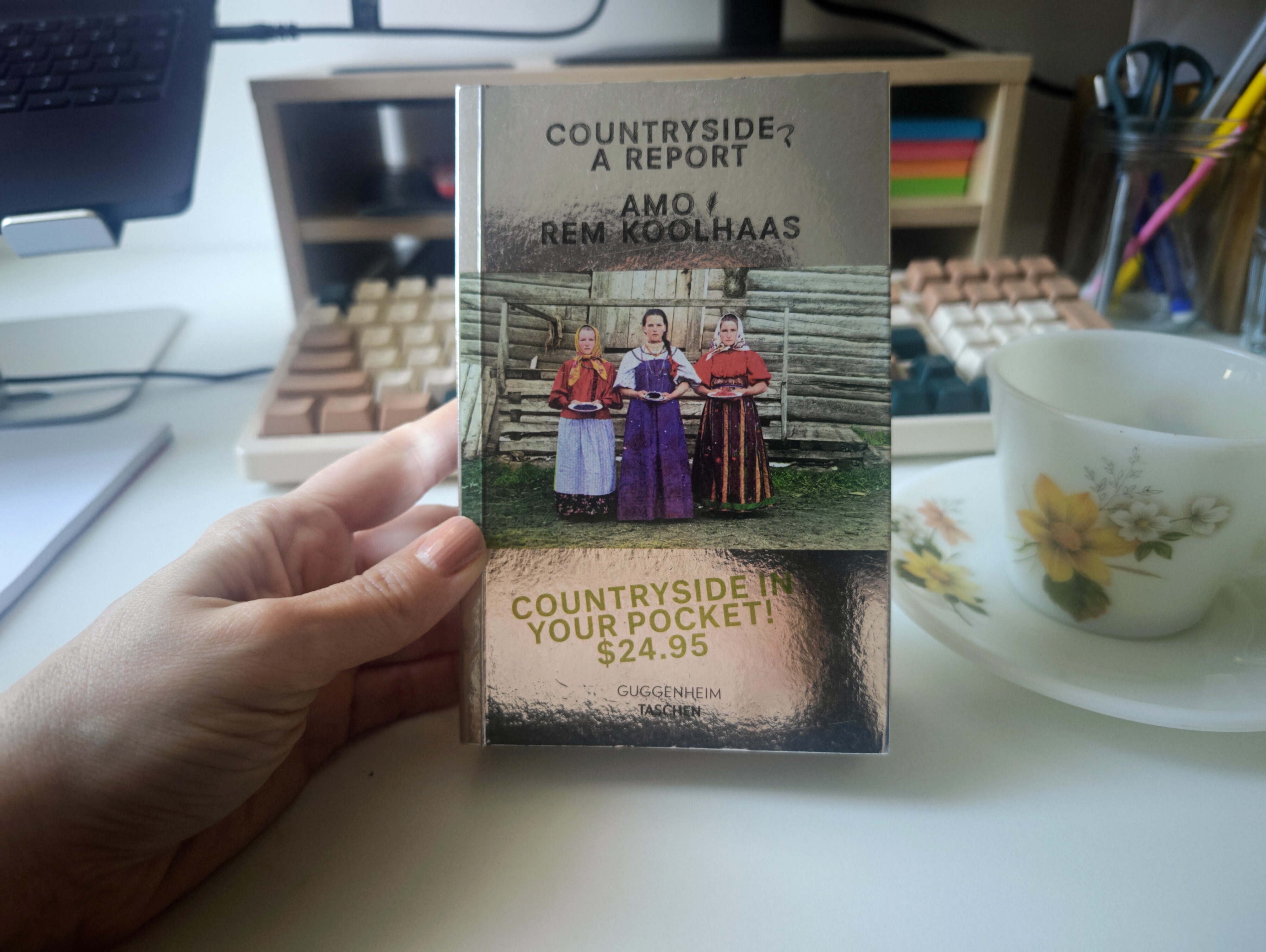 Countryside: A Report by AMO and Rem Koolhaas, held in front of a desk with a mechanical keyboard and a teacup.